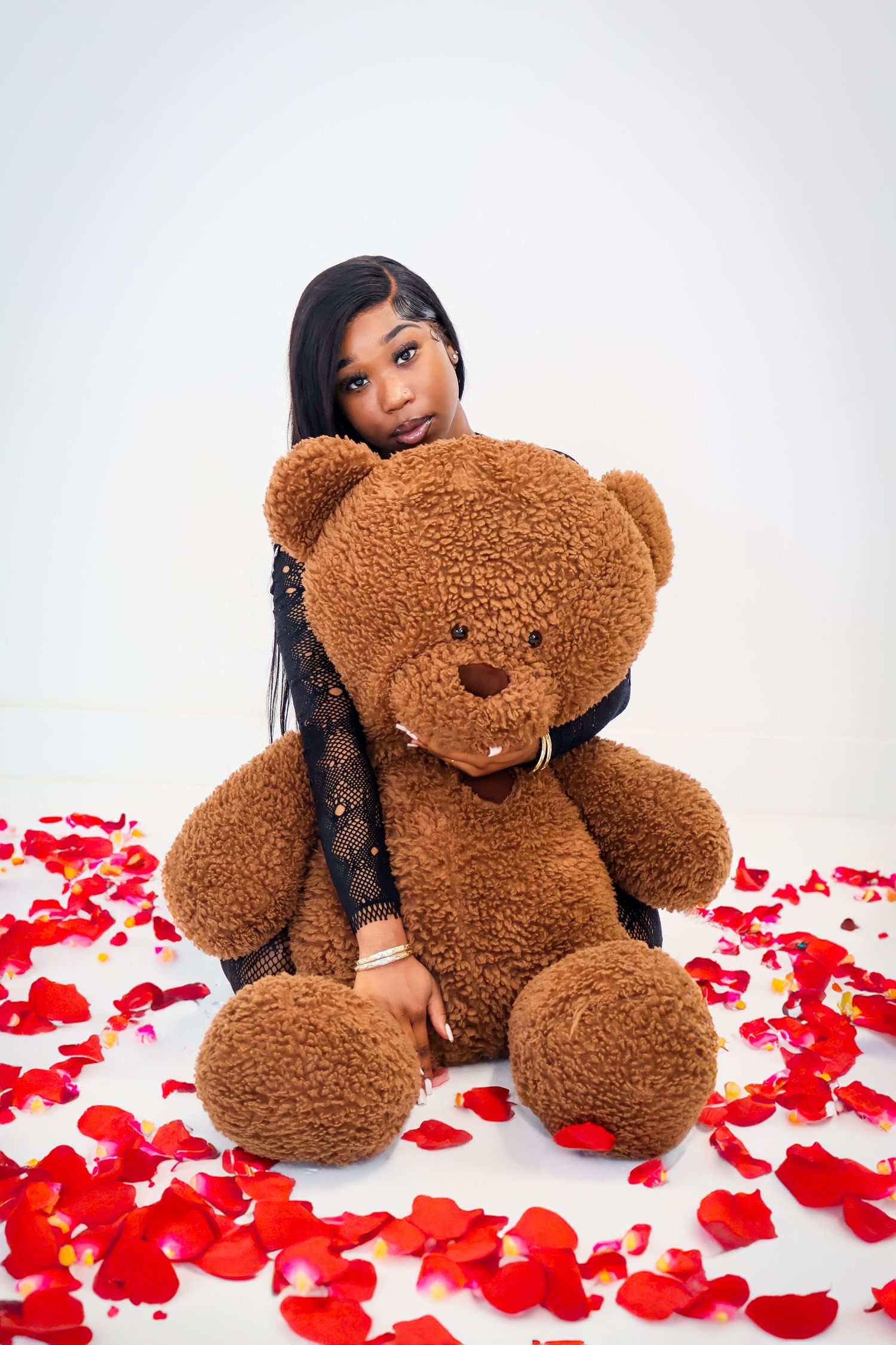 Young woman hugging a large brown teddy bear surrounded by red rose petals on white floor.