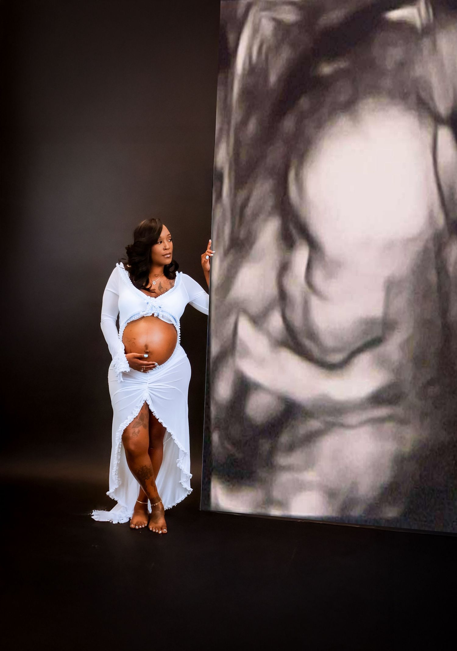 Pregnant woman in white dress poses beside large ultrasound image backdrop in elegant maternity photo shoot.