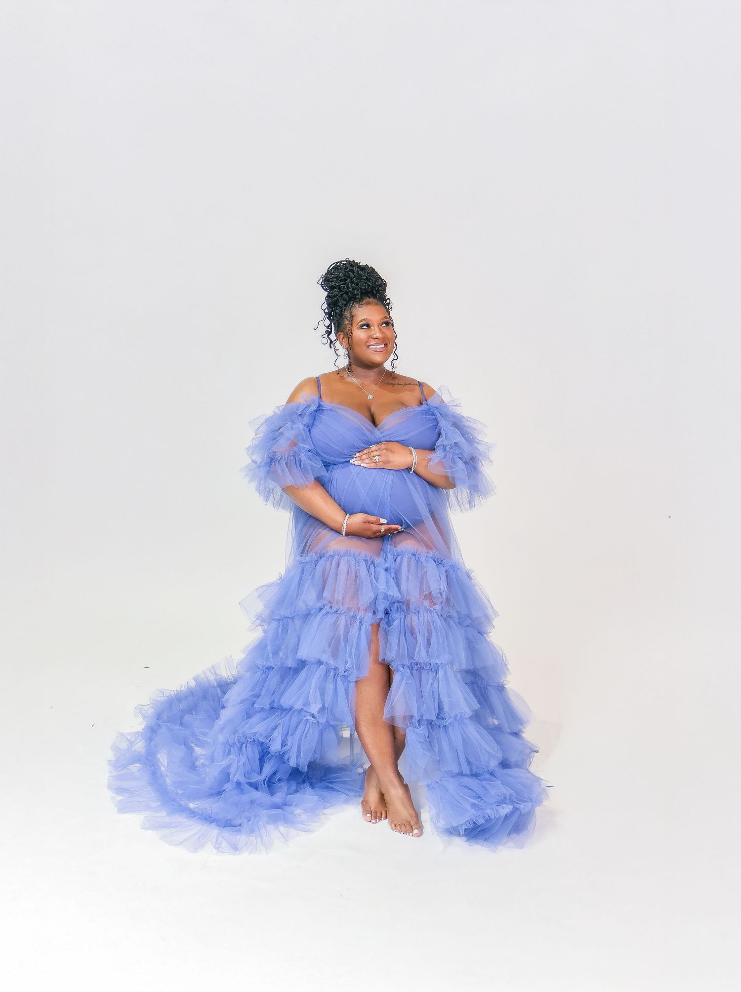 Pregnant woman in a flowing periwinkle blue ruffled tulle maternity gown posing gracefully in a studio.