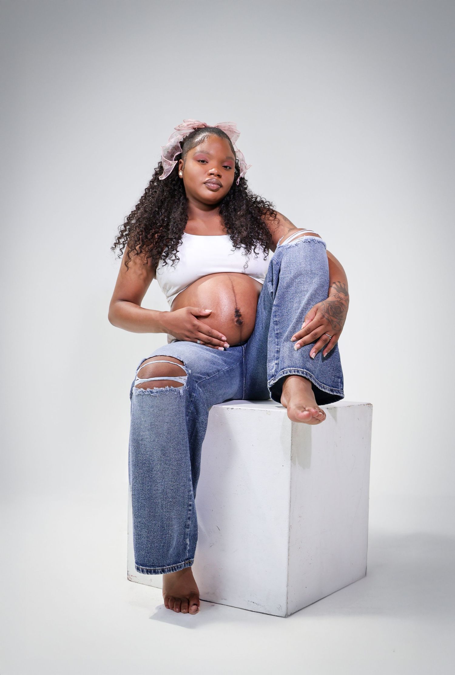 Pregnant Black woman sitting on white cube, wearing ripped jeans and white crop top, cradling baby bump, curly hair with pink bow.