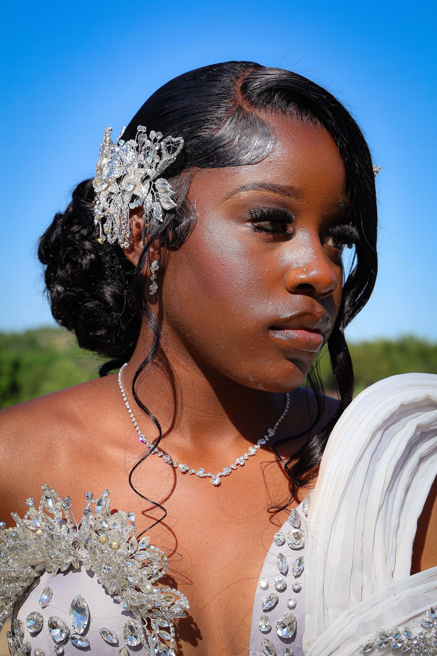 Young Black woman in elegant beaded gown with crystal hair accessory and diamond necklace poses outdoors under blue sky.