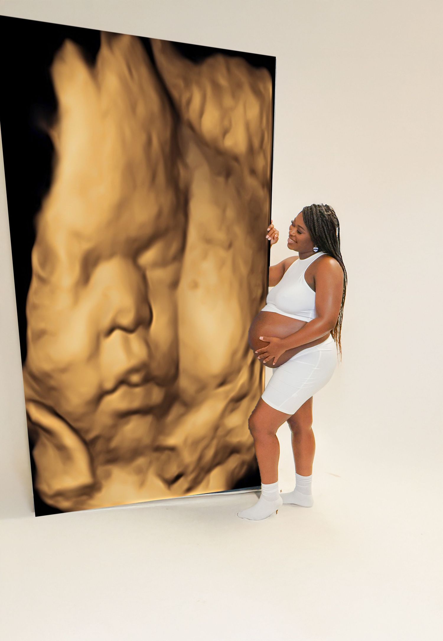 Pregnant woman in white outfit poses beside a large 3D ultrasound image of her unborn baby.