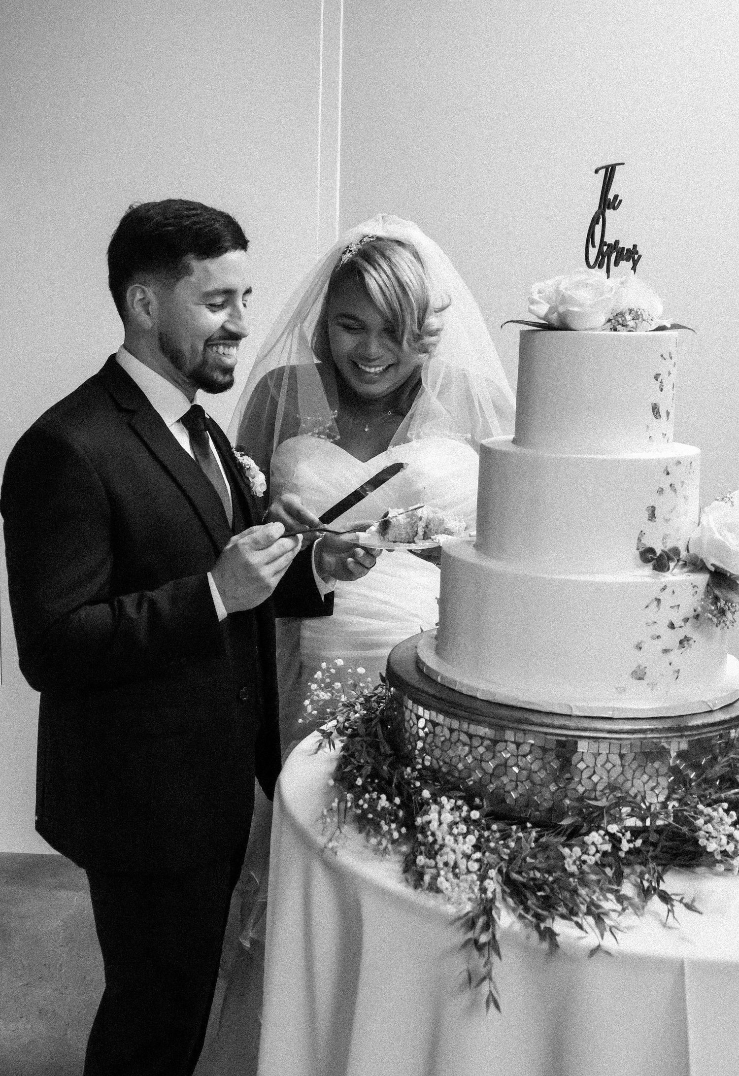 Black and white photo of smiling couple cutting tiered wedding cake at their reception.