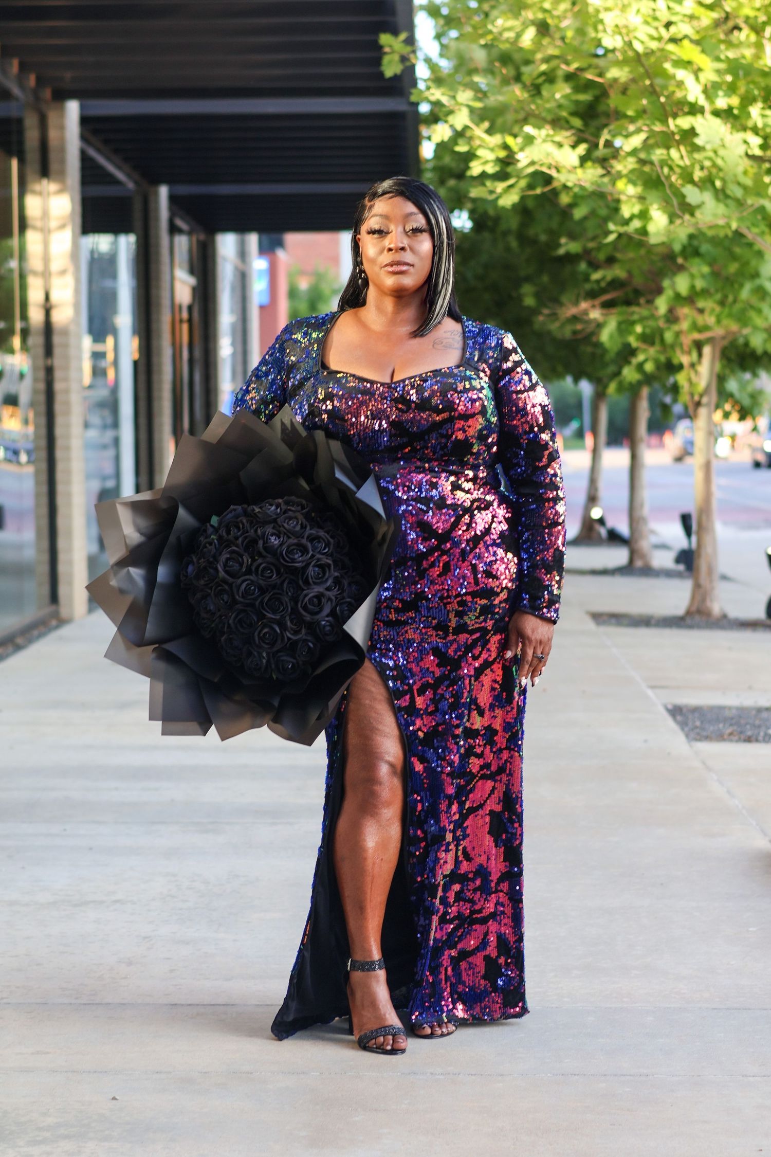 A woman in a stunning iridescent sequined gown holds black roses while posing on a city sidewalk.
