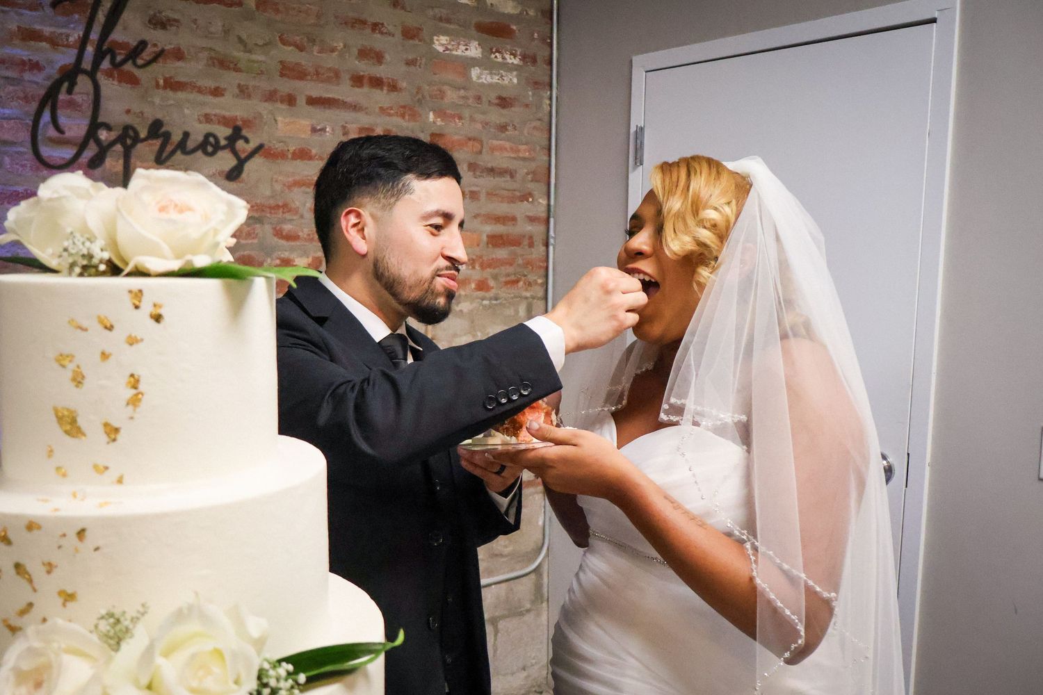 Bride and groom feeding each other wedding cake at their reception, with a white floral tiered cake nearby.