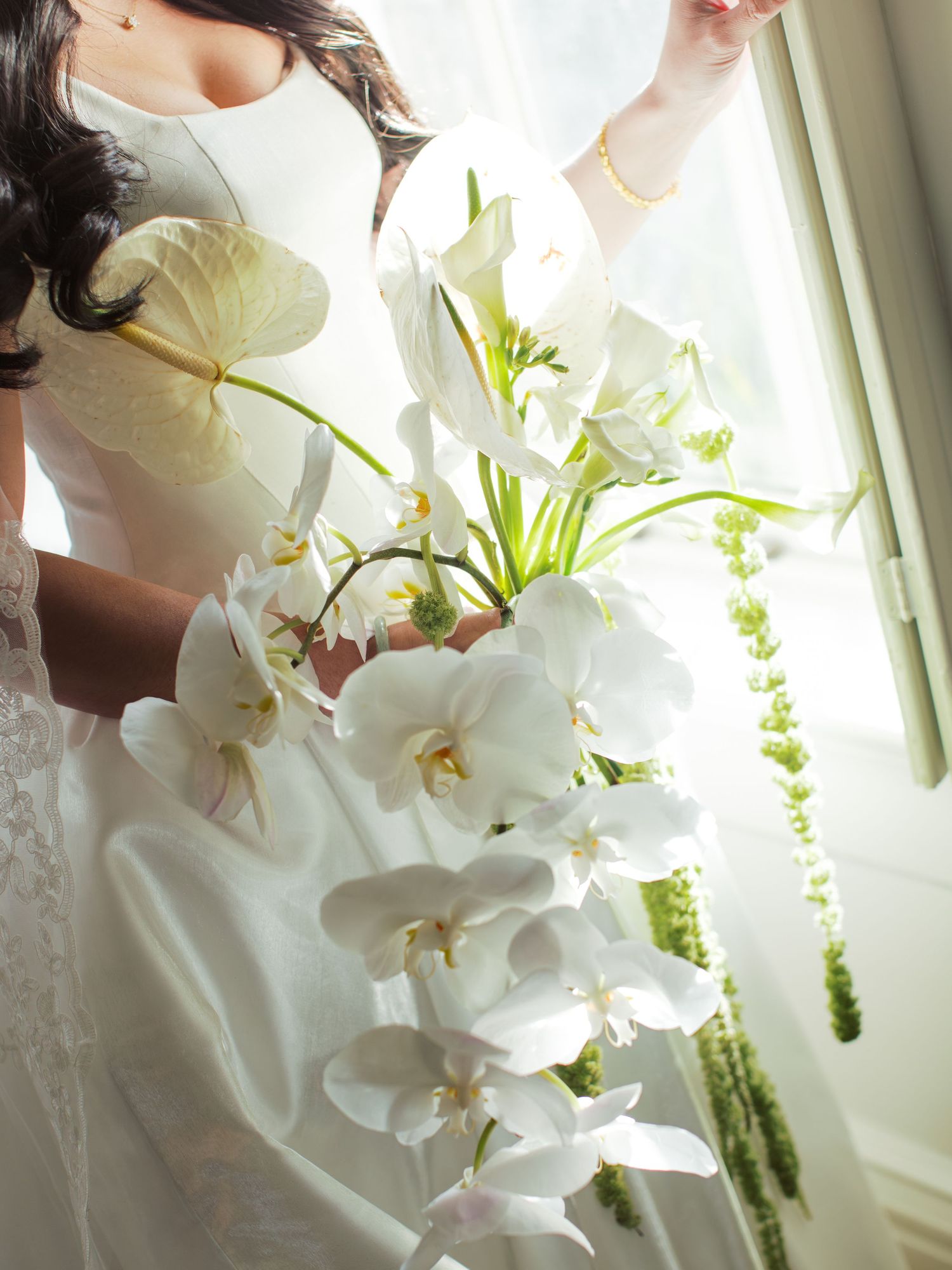 Bride in white wedding dress holding elegant cascading bouquet of white orchids and lilies near a sunlit window.