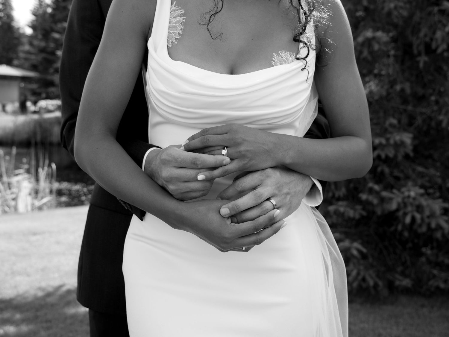 Black and white wedding photo of bride in white gown with groom embracing her from behind, showing wedding rings.