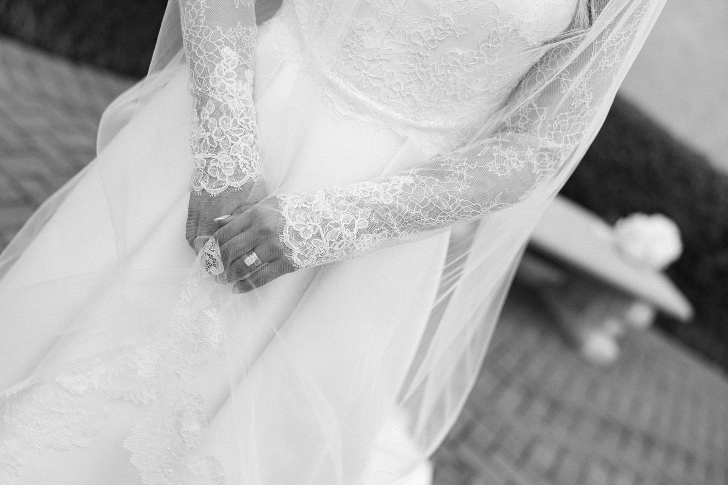 Black and white photo of a bride in a lace wedding dress and veil, hands clasped together wearing a ring.
