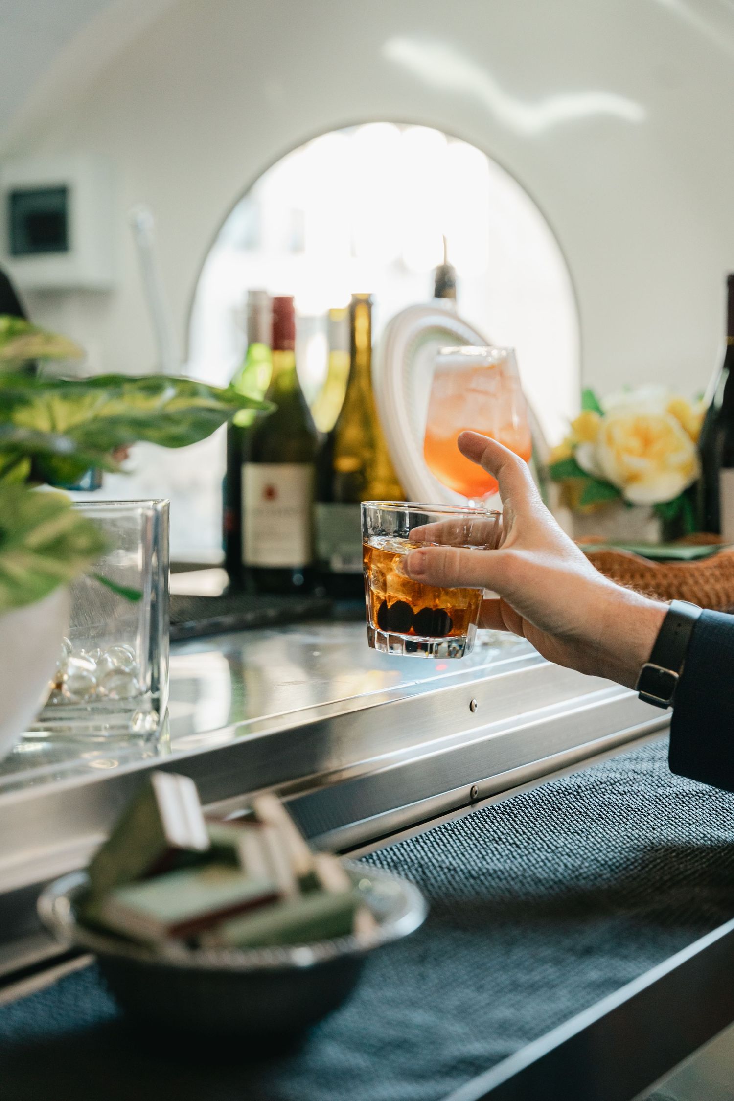Hand holding a cocktail glass with wine bottles and flowers in the background.