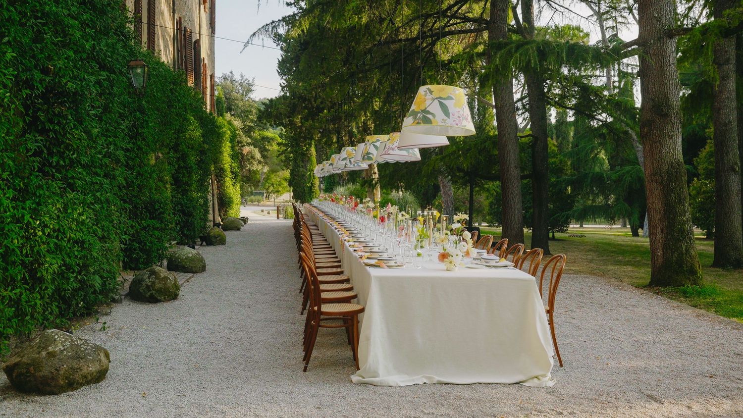 Elegant outdoor wedding reception with long white-draped table, wooden chairs, and string lights in a lush garden setting.