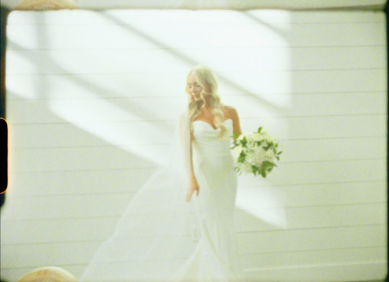 Blonde bride in a white strapless mermaid wedding gown holding a bouquet of white flowers in a sunlit room.