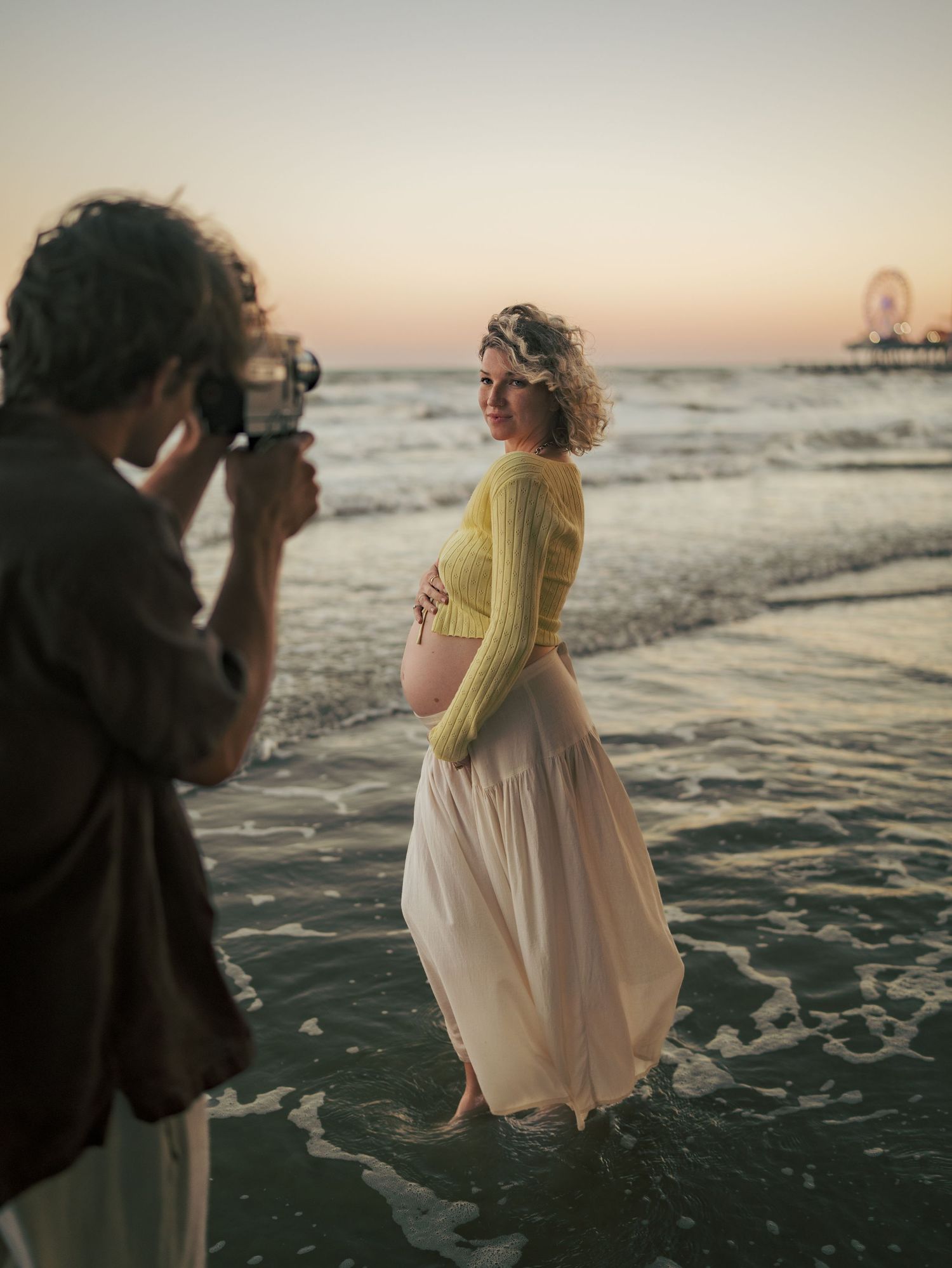 Pregnant woman in yellow crop top and pink skirt poses on beach at sunset while photographer captures her maternity photo.