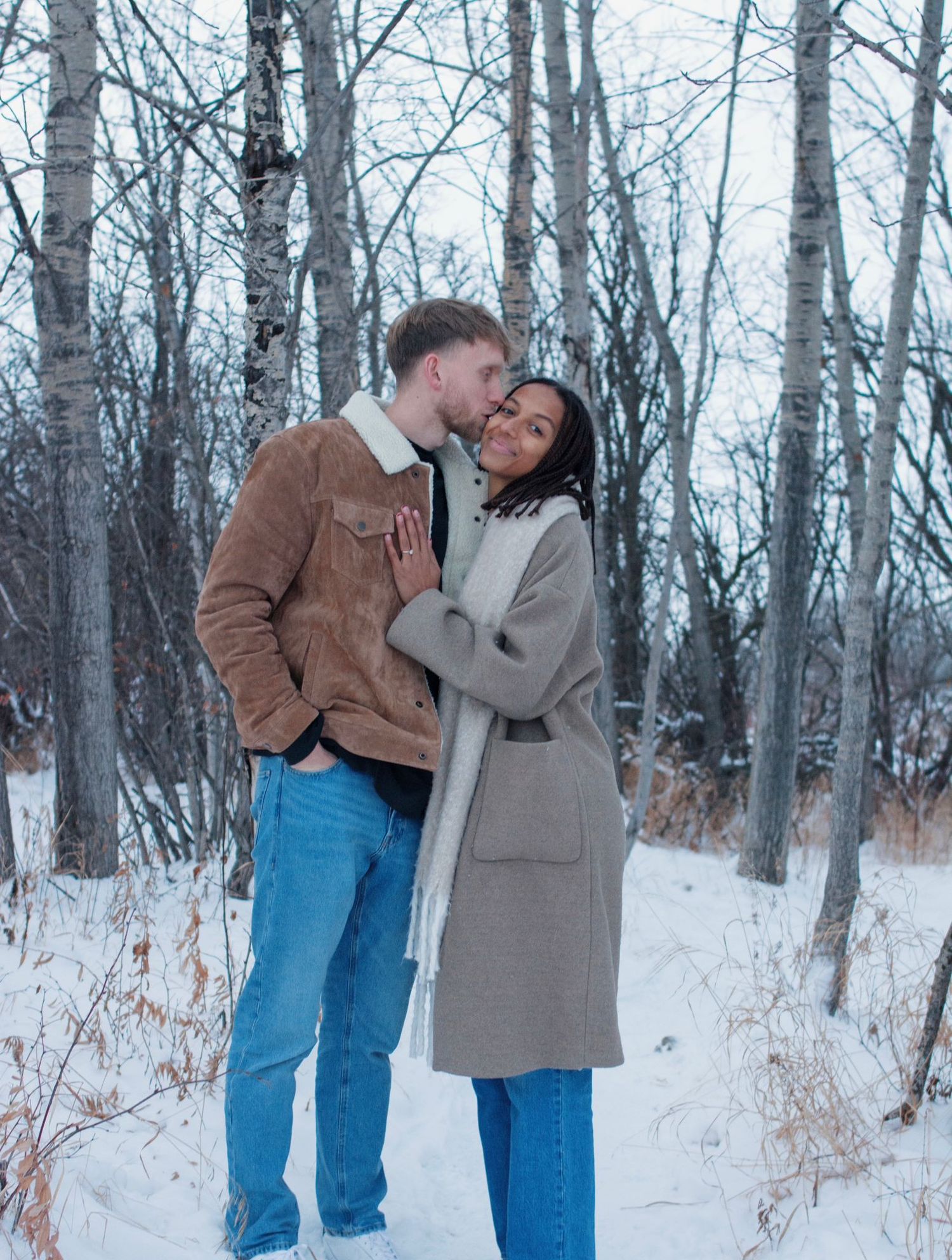 A couple shares a tender moment together in a snowy winter forest setting wearing coats and jeans.