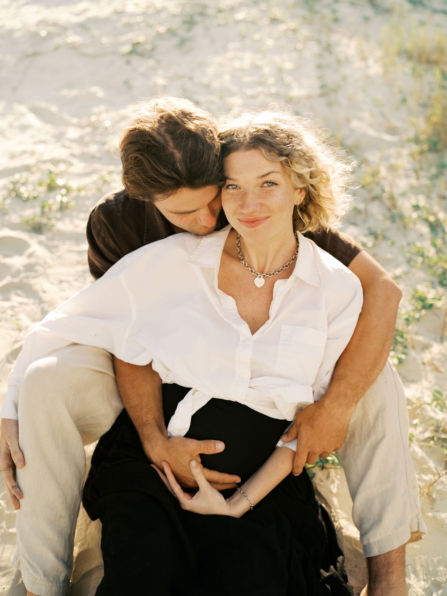 Couple sharing a tender maternity beach moment, woman in white shirt cradling baby bump while partner embraces her from behind.