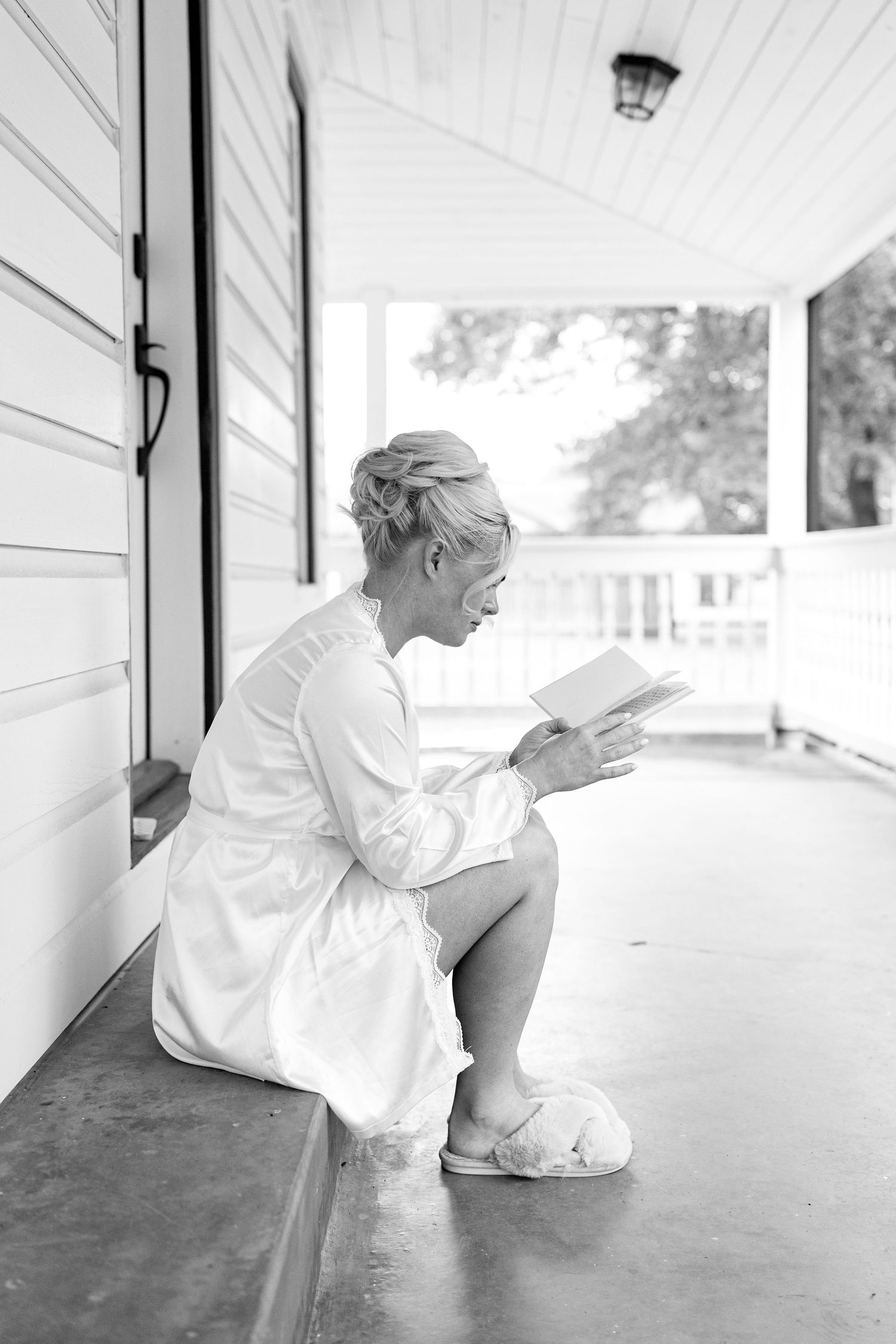 Black and white photo of a bride in a white robe and slippers reading a letter on a porch before her wedding.