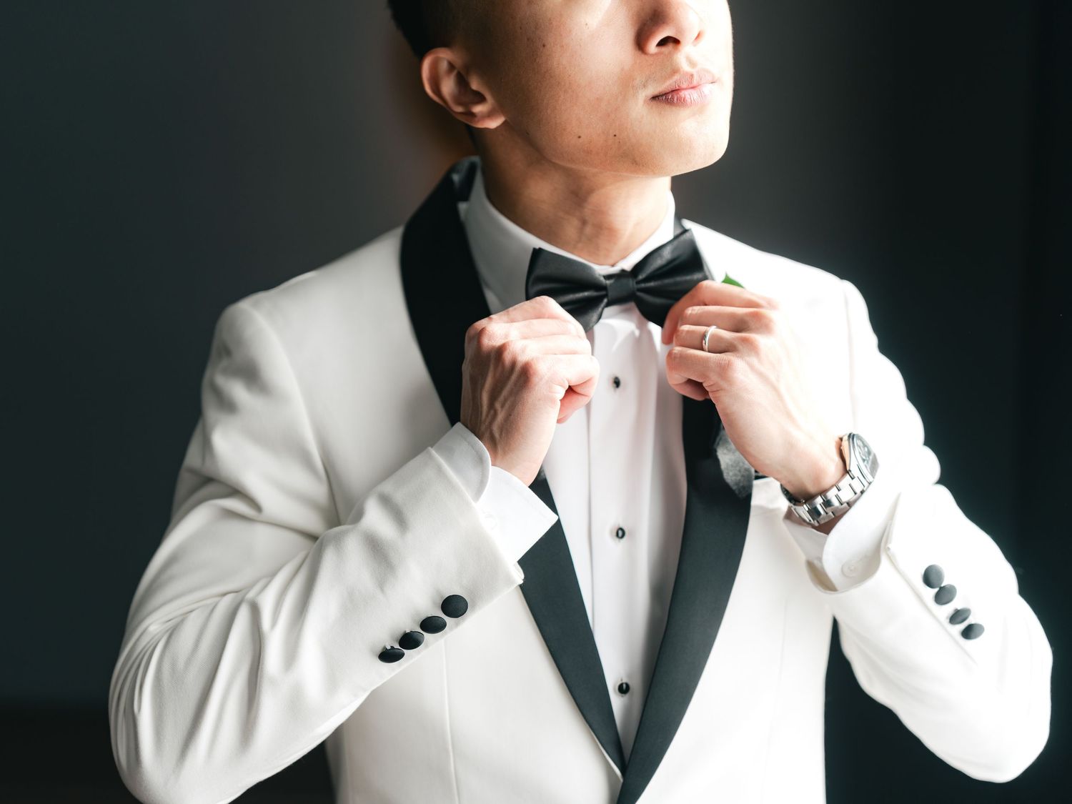 Man in white tuxedo with black lapels adjusting bow tie, wearing a watch, formal wedding attire.