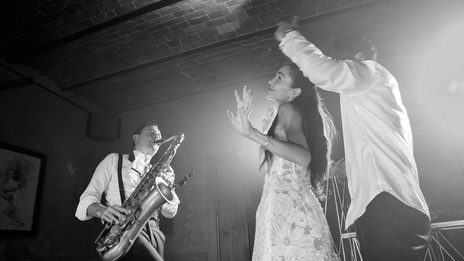 Black and white photo of a saxophonist playing beside a woman in a flowing dress dancing on stage with a man in a light shirt.