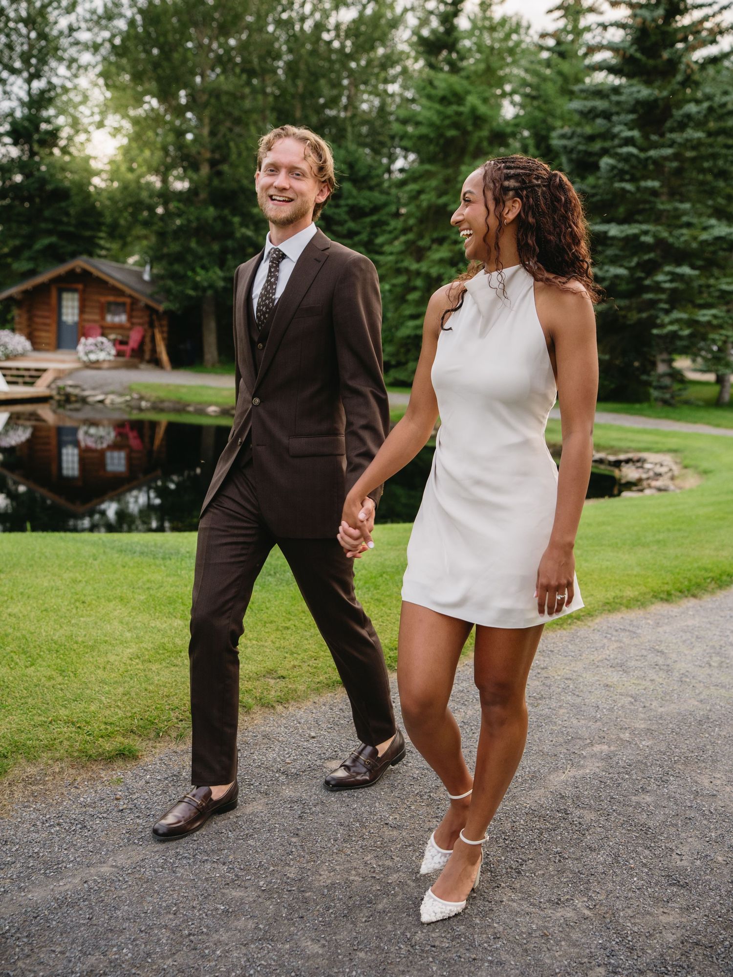 Couple in wedding attire holding hands, man in dark suit, woman in white mini dress, walking by a pond.