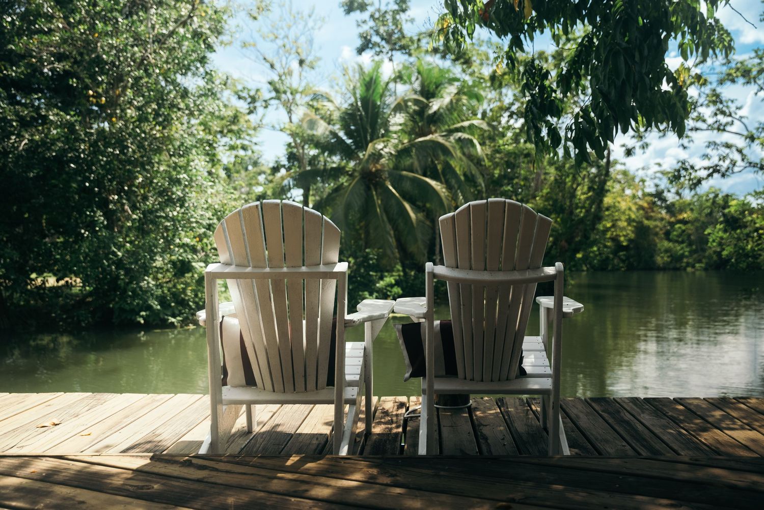 Two white Adirondack chairs on a wooden deck overlooking a tropical waterfront with palm trees and lush vegetation.
