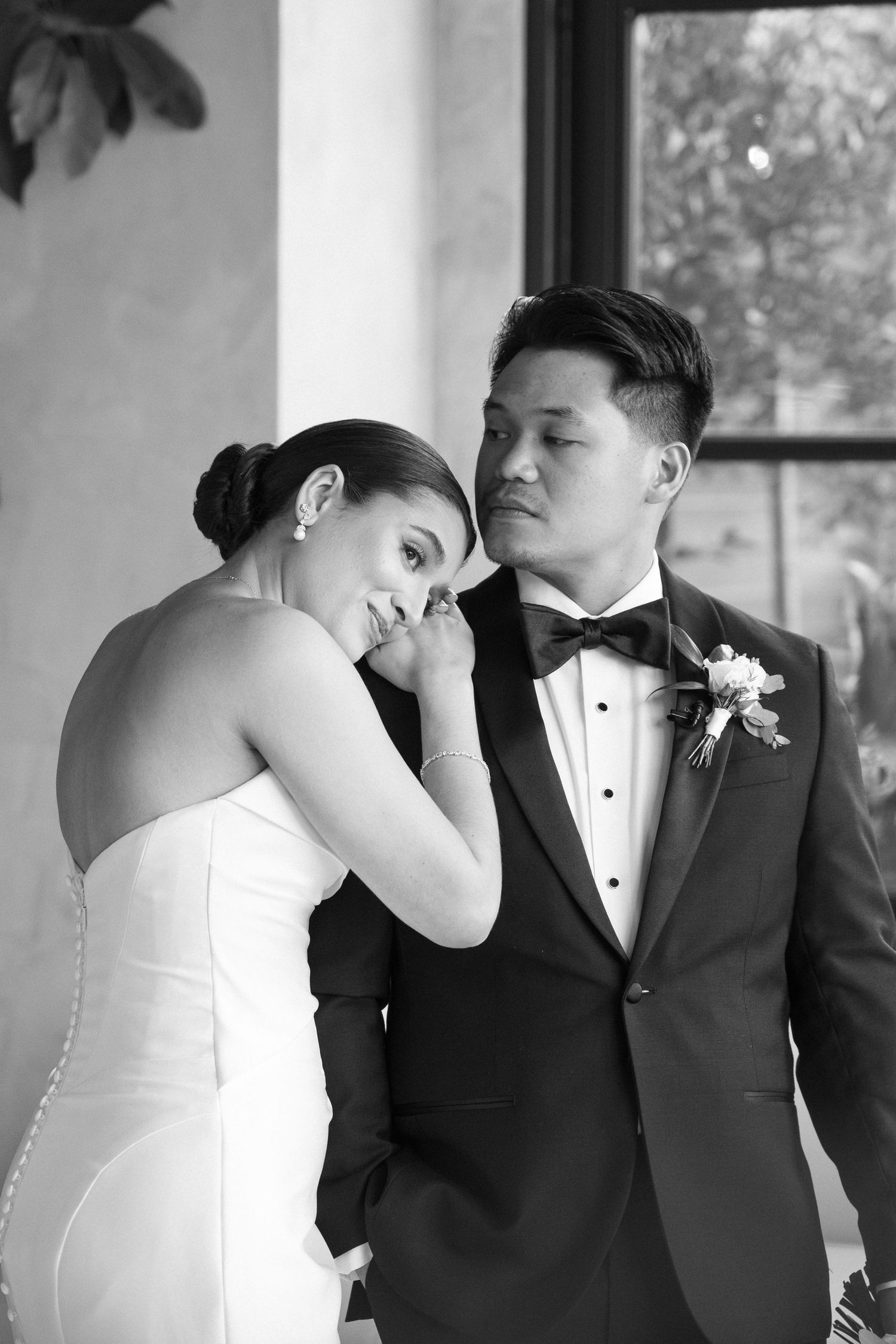Black and white wedding photo of bride leaning head on groom's shoulder in elegant tuxedo with boutonniere.