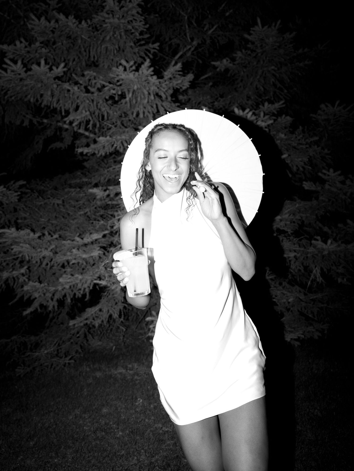 Black and white photo of a smiling woman in a white dress holding a drink, standing outdoors at night near pine trees.