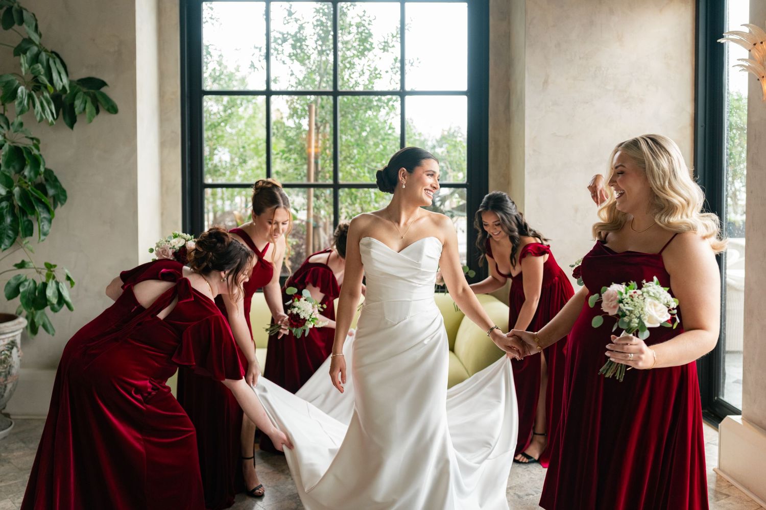 Bride in white gown surrounded by bridesmaids in red velvet dresses helping adjust her train indoors.