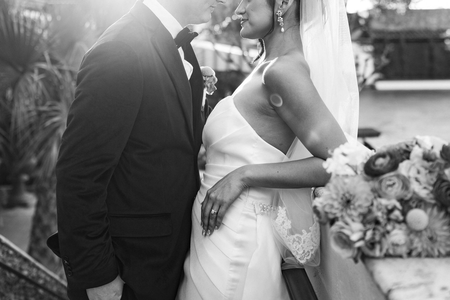 Black and white wedding photo of bride in lace gown and groom in tuxedo embracing outdoors.