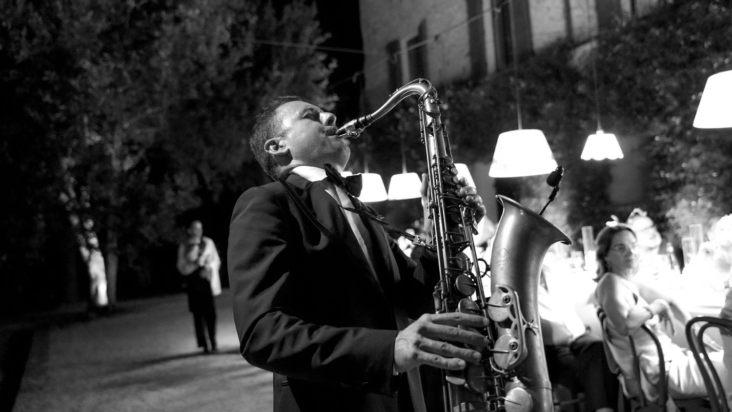 Black and white photo of a jazz musician playing saxophone outdoors at a night event.