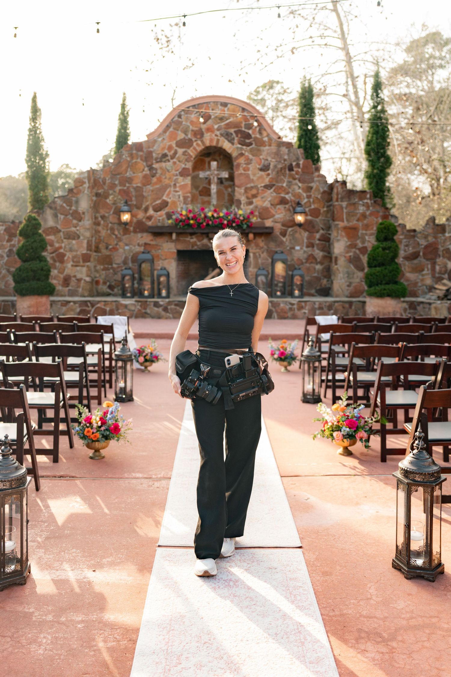 Woman in black outfit walks down outdoor wedding aisle at stone chapel venue with floral arrangements.
