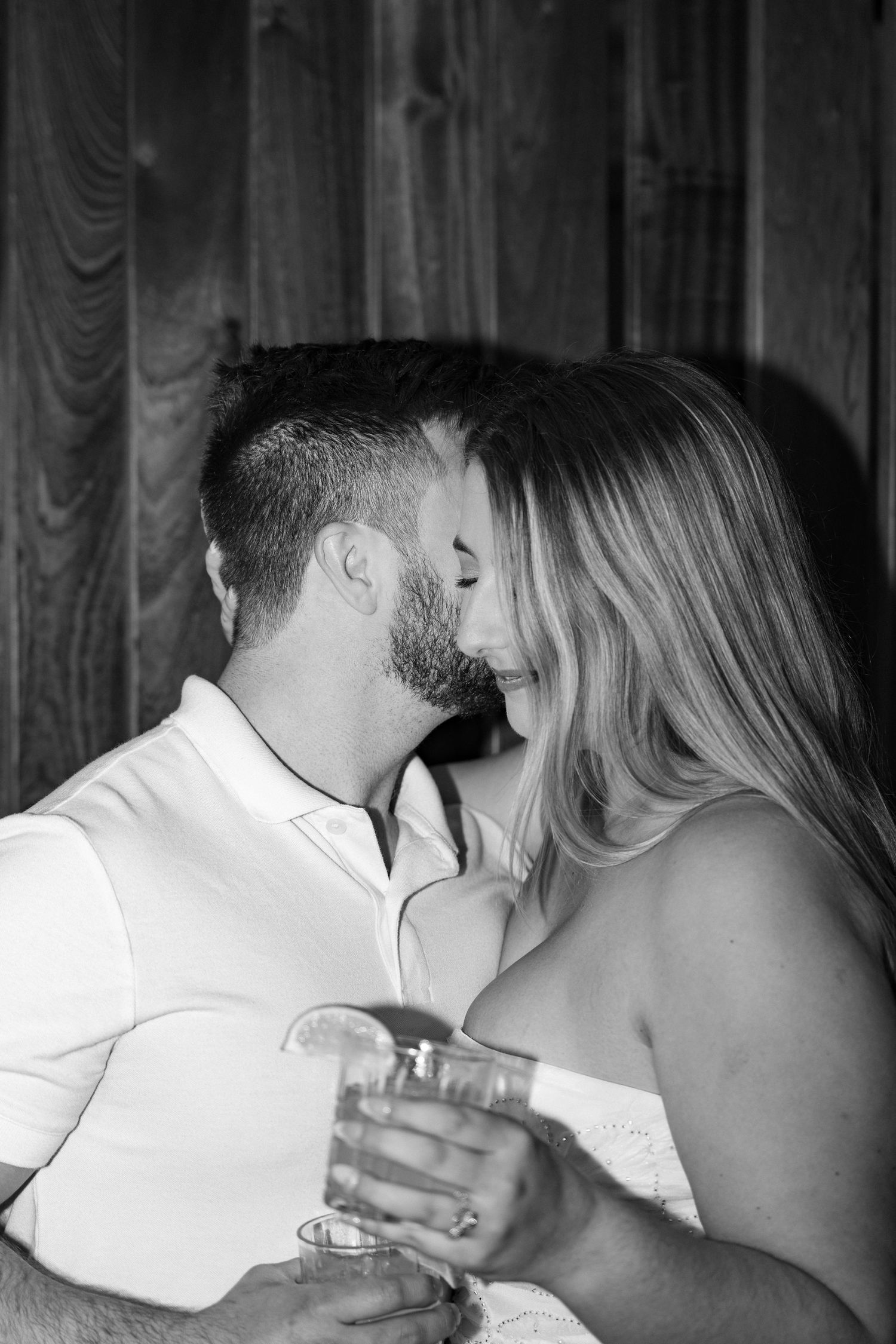 Black and white photo of a couple sharing a romantic kiss while holding cocktail glasses against a wooden backdrop.