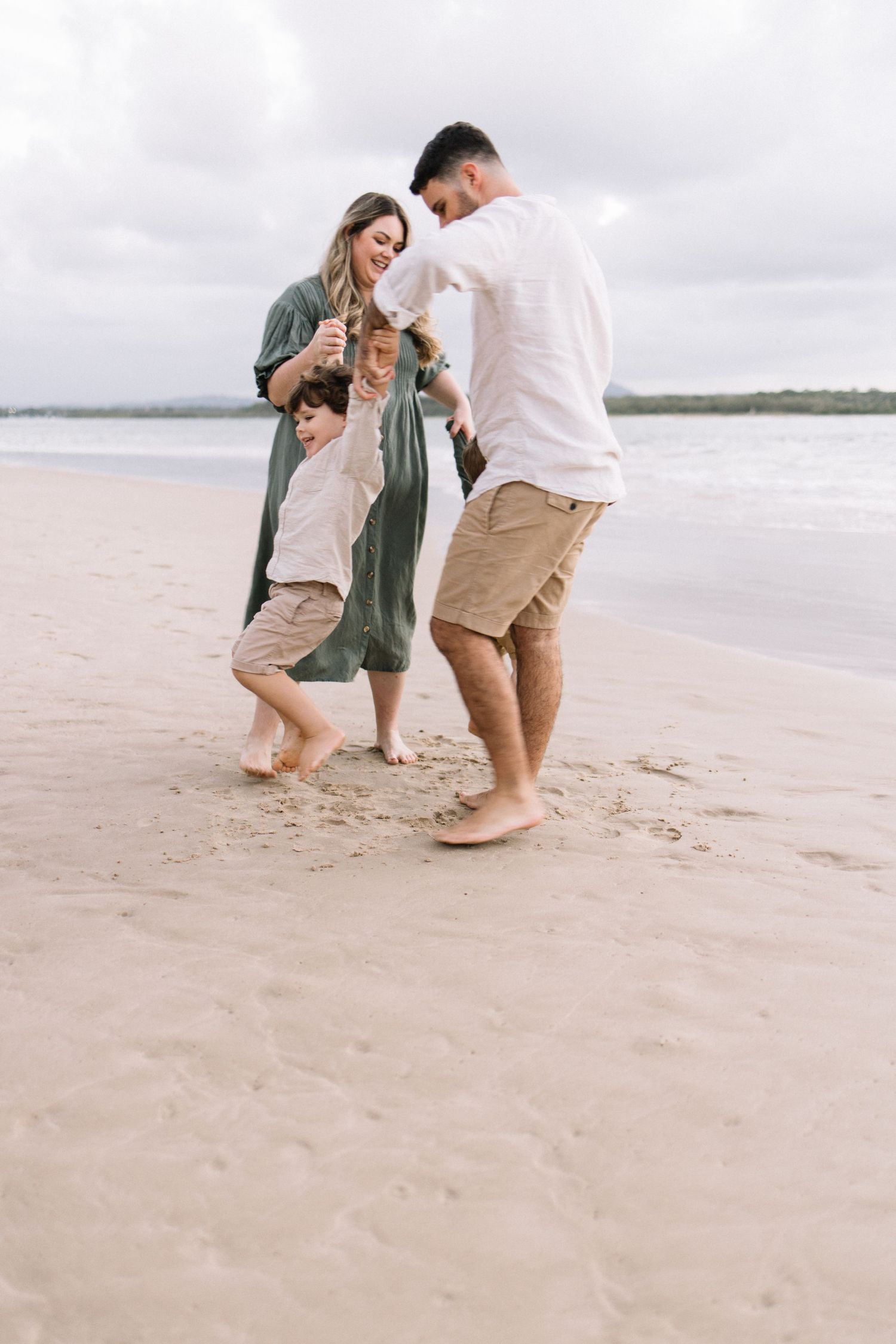A happy family plays together on a sandy beach, swinging their young child between them.