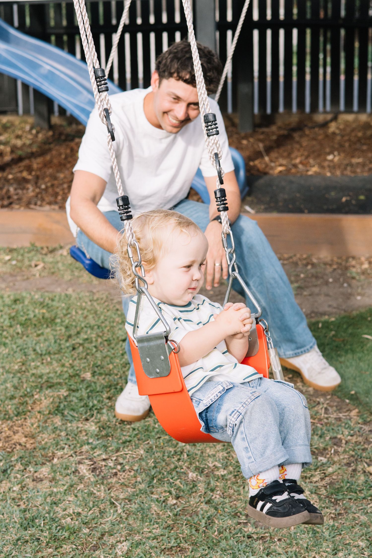 Father smiling while pushing toddler on orange baby swing at outdoor playground on sunny day.