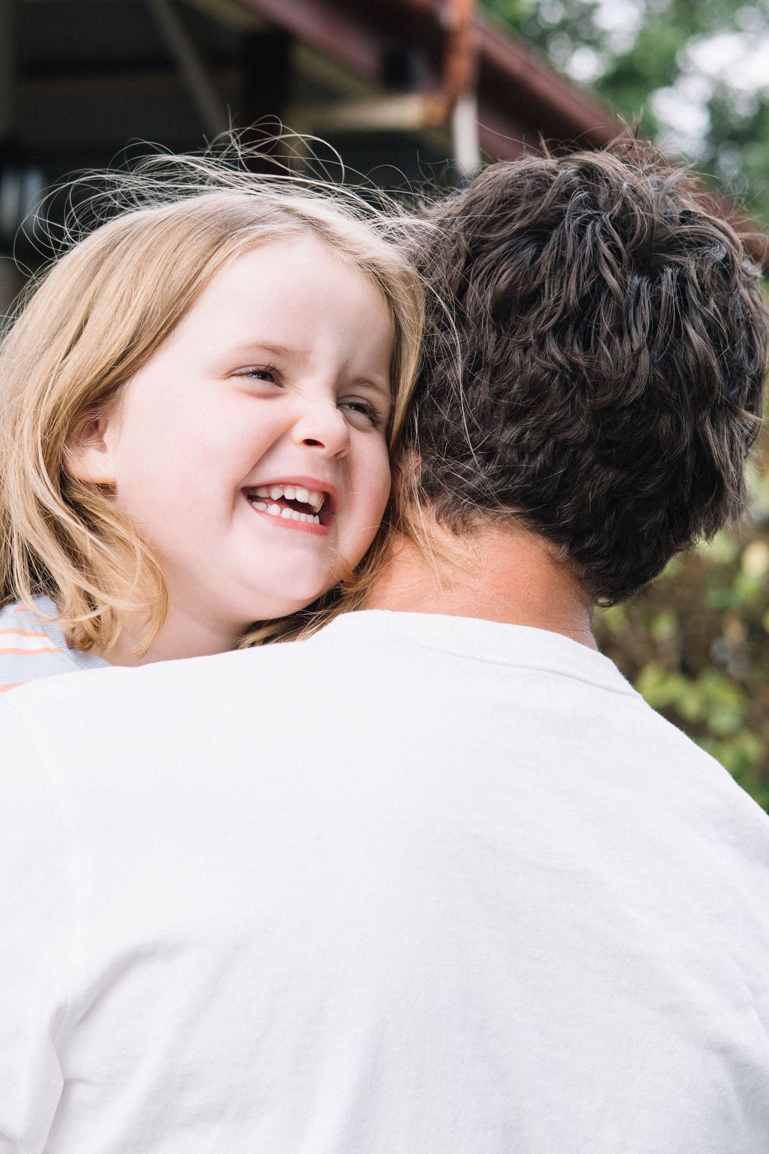 A laughing young girl with blonde hair being held by an adult in a white shirt outdoors.