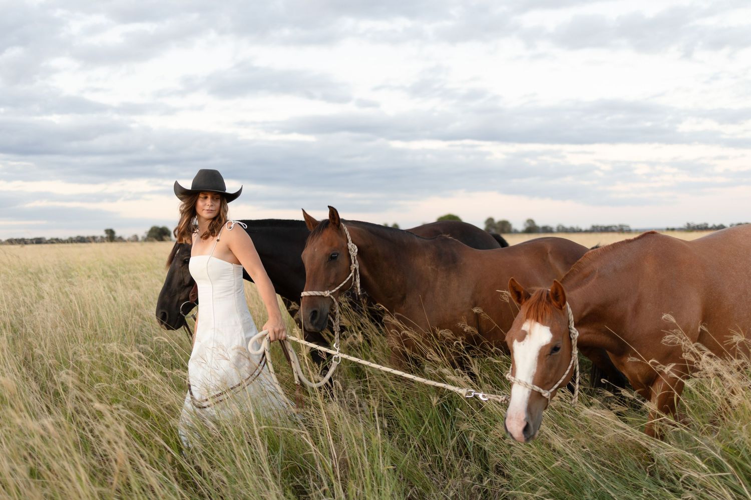Woman in white dress and cowboy hat leading two horses through tall grass in a rural field.