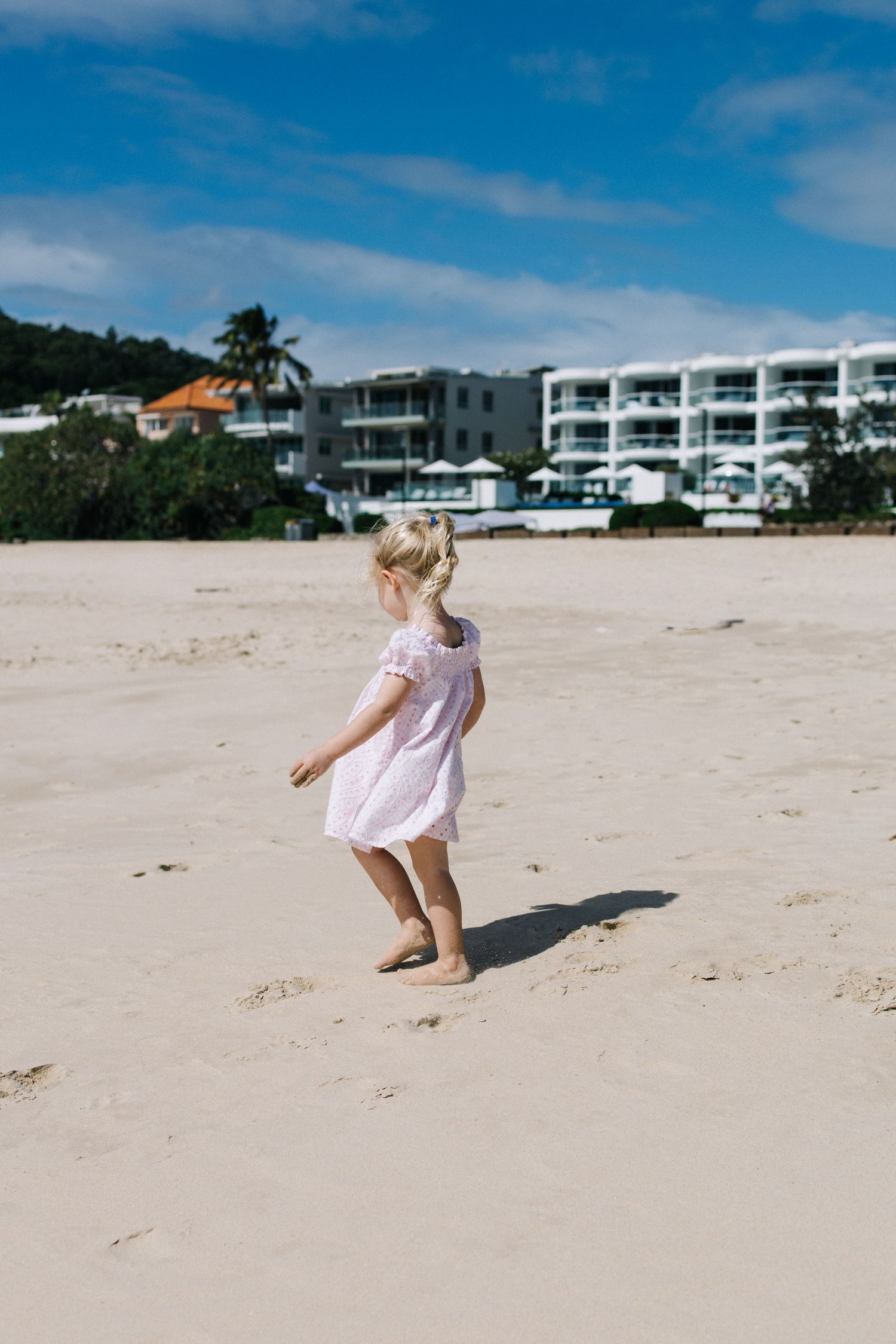 Young blonde girl in pink dress walking barefoot on white sand beach with resort buildings and palm trees in background.