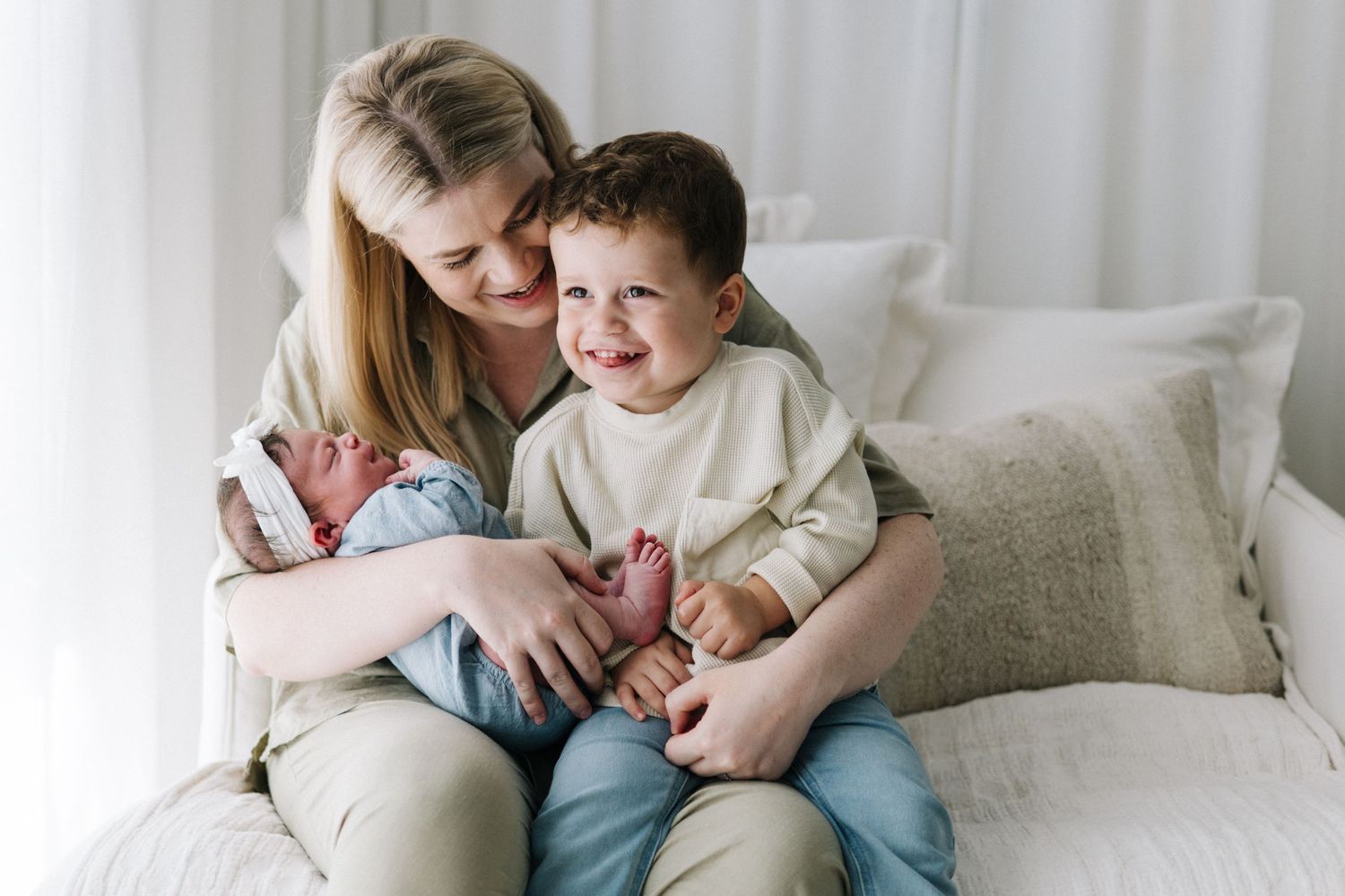 A smiling mother holds her newborn baby while sitting with her happy toddler son on a white couch.