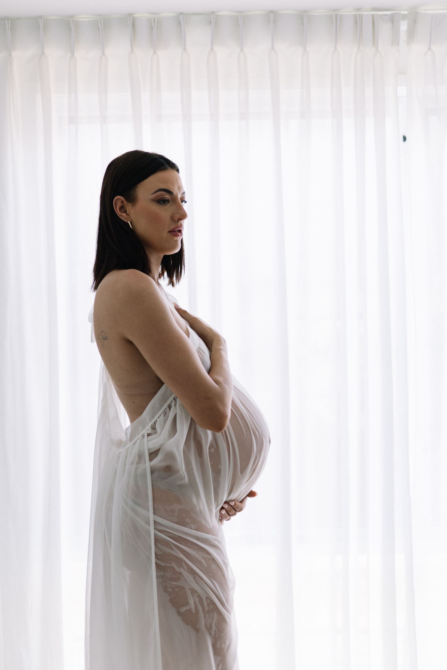 Pregnant woman in white sheer lace robe holding belly sideways against bright curtains.