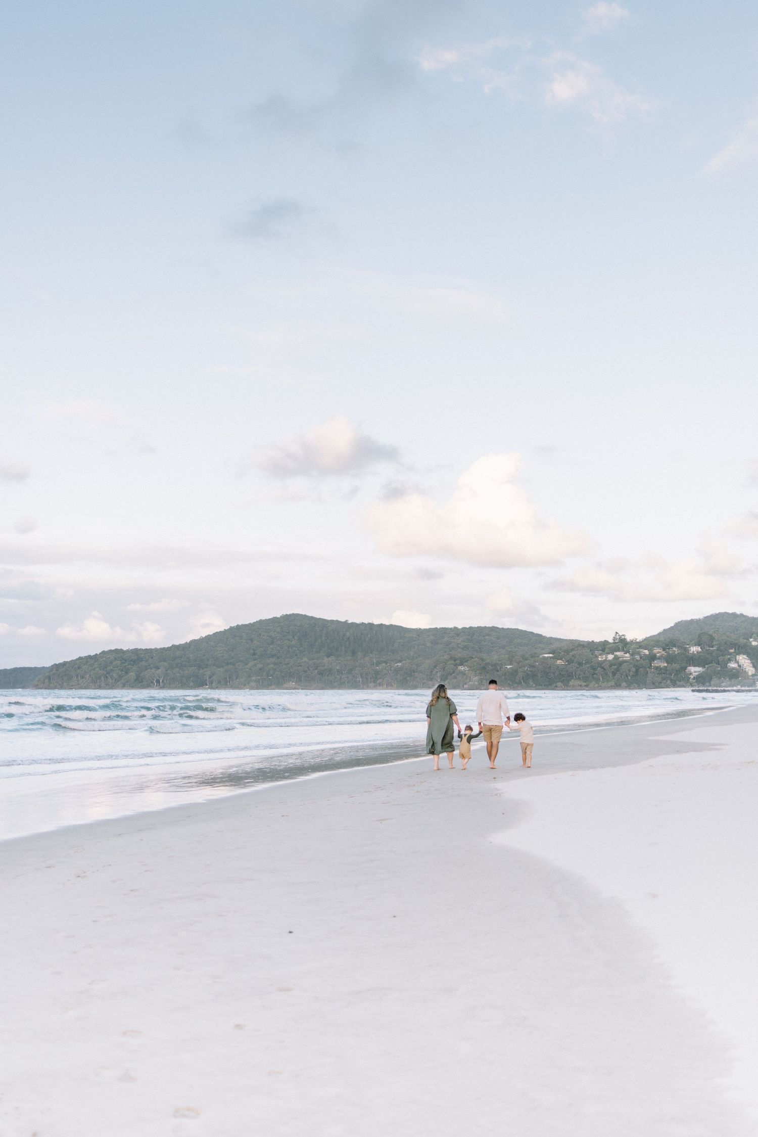 Family walking along a white sandy beach at sunset with calm ocean waves and green hills in the background.