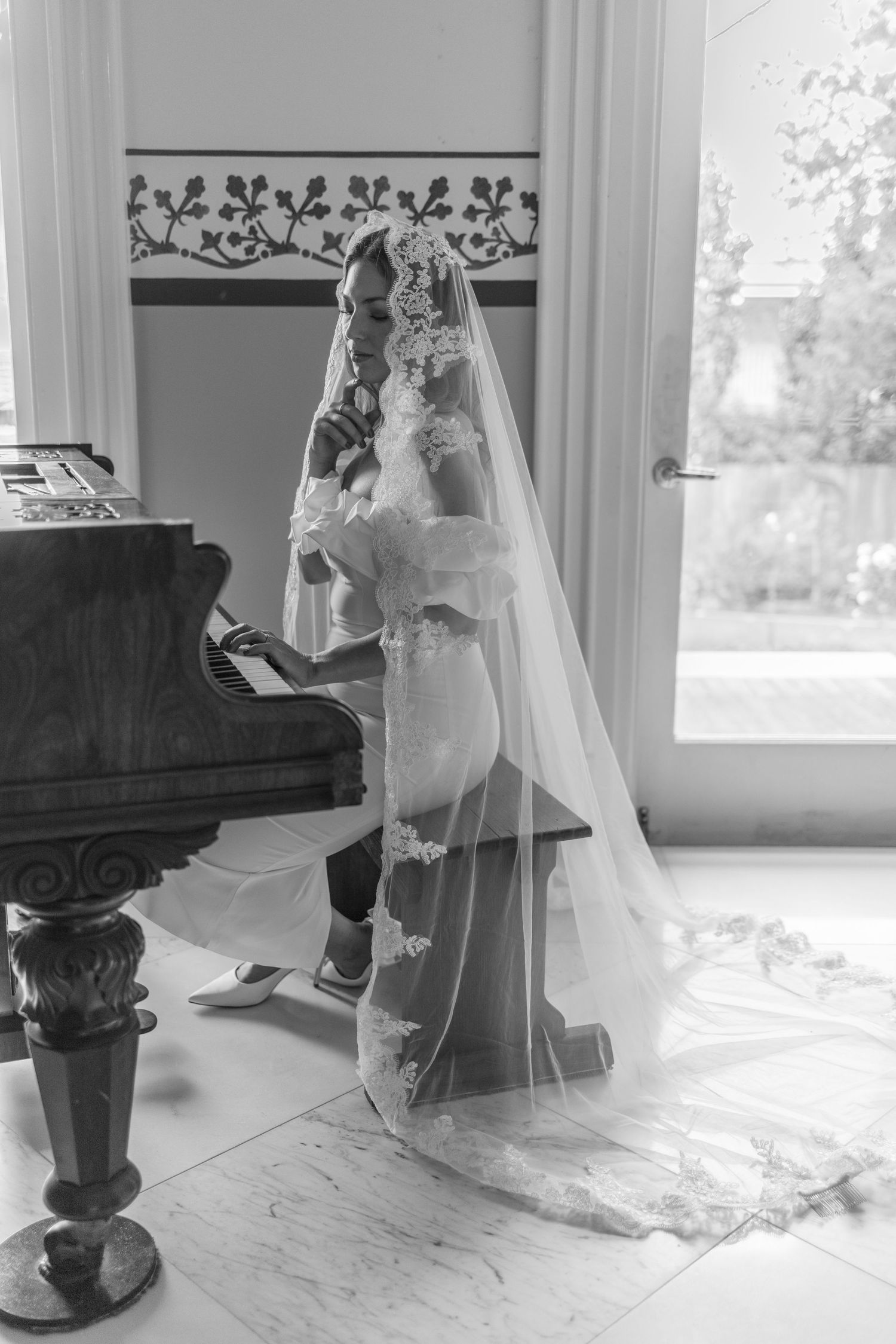 Bride in lace veil playing grand piano in black and white wedding photo near bright window.