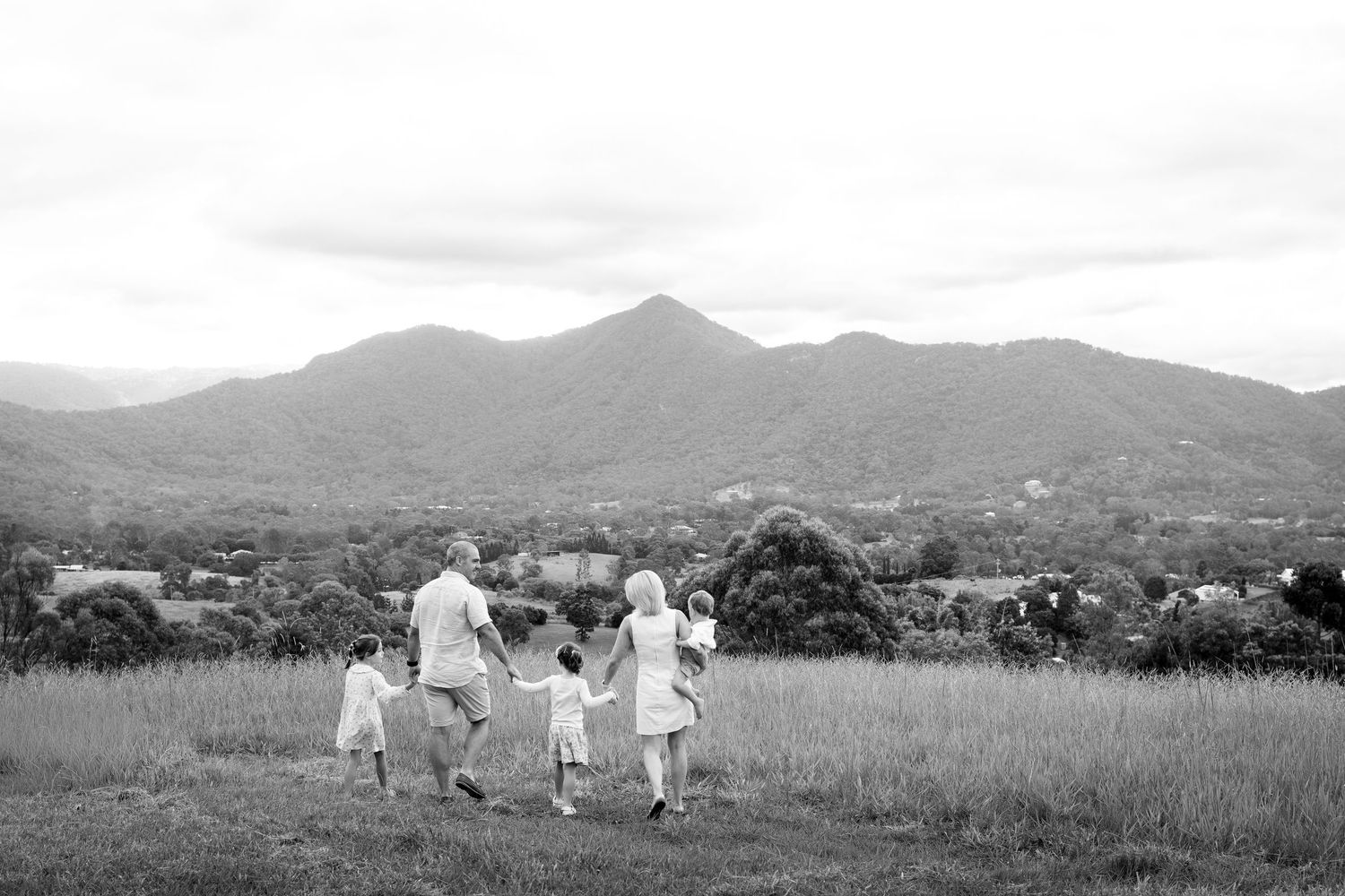 Black and white photo of a family walking through a field holding hands with mountains in the background.
