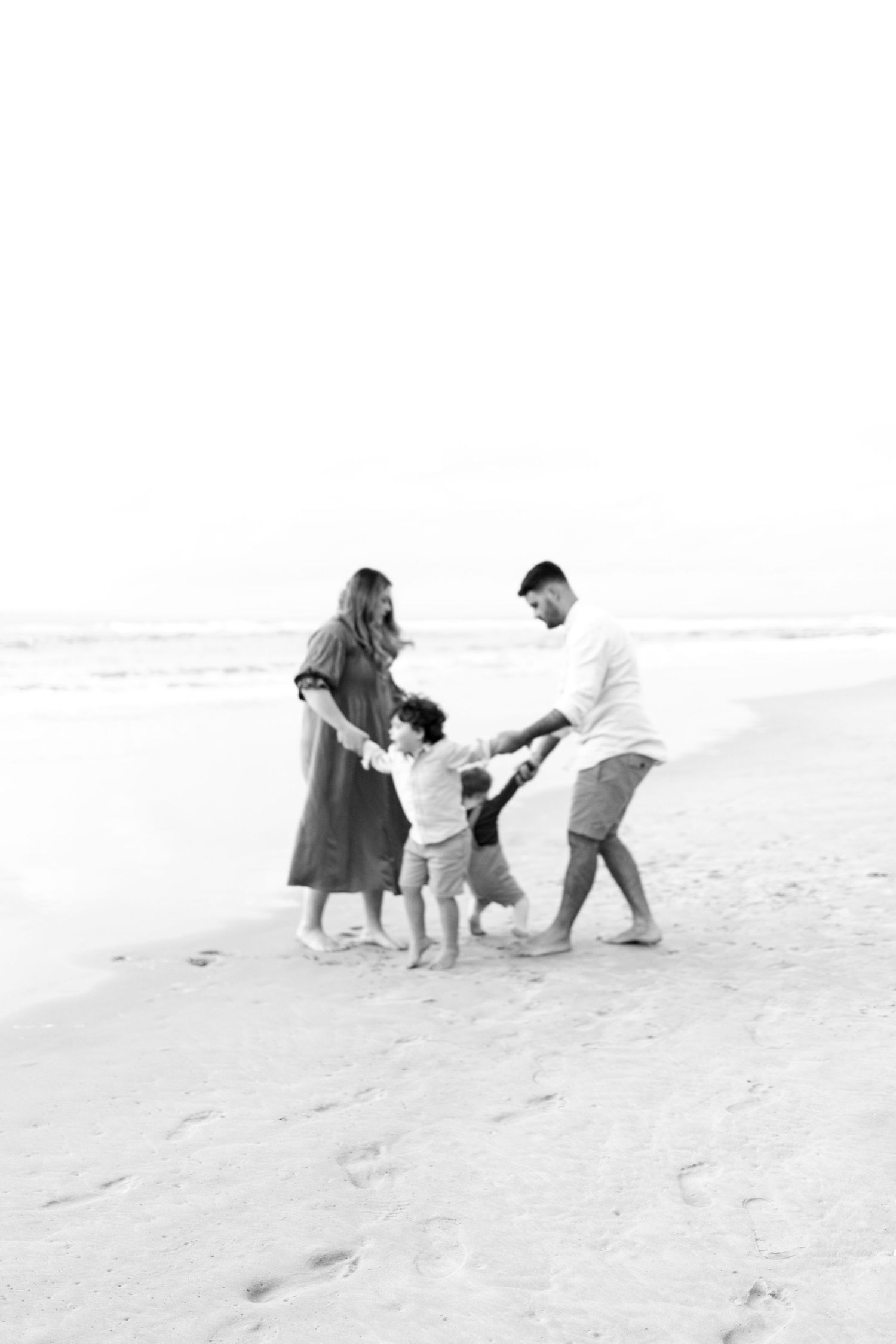 Black and white photo of a family of four playing together on a sandy beach near the ocean.