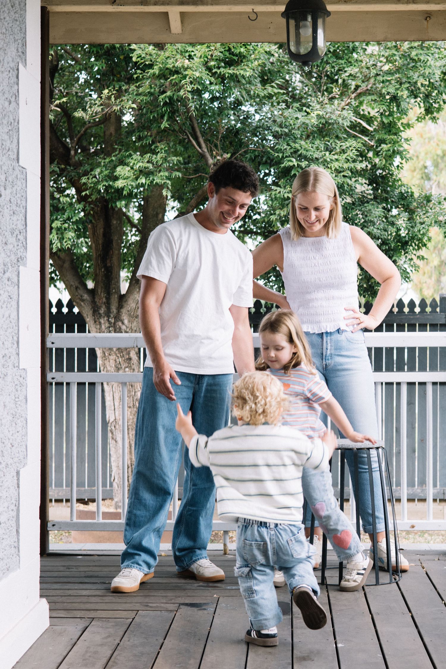 Family of four smiling on a wooden porch, parents in white tees and jeans watching two young children play.