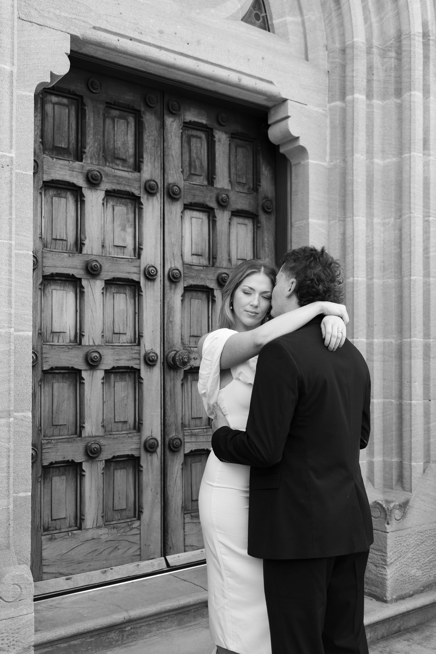 Black and white photo of a bride and groom embracing in front of ornate wooden church doors.