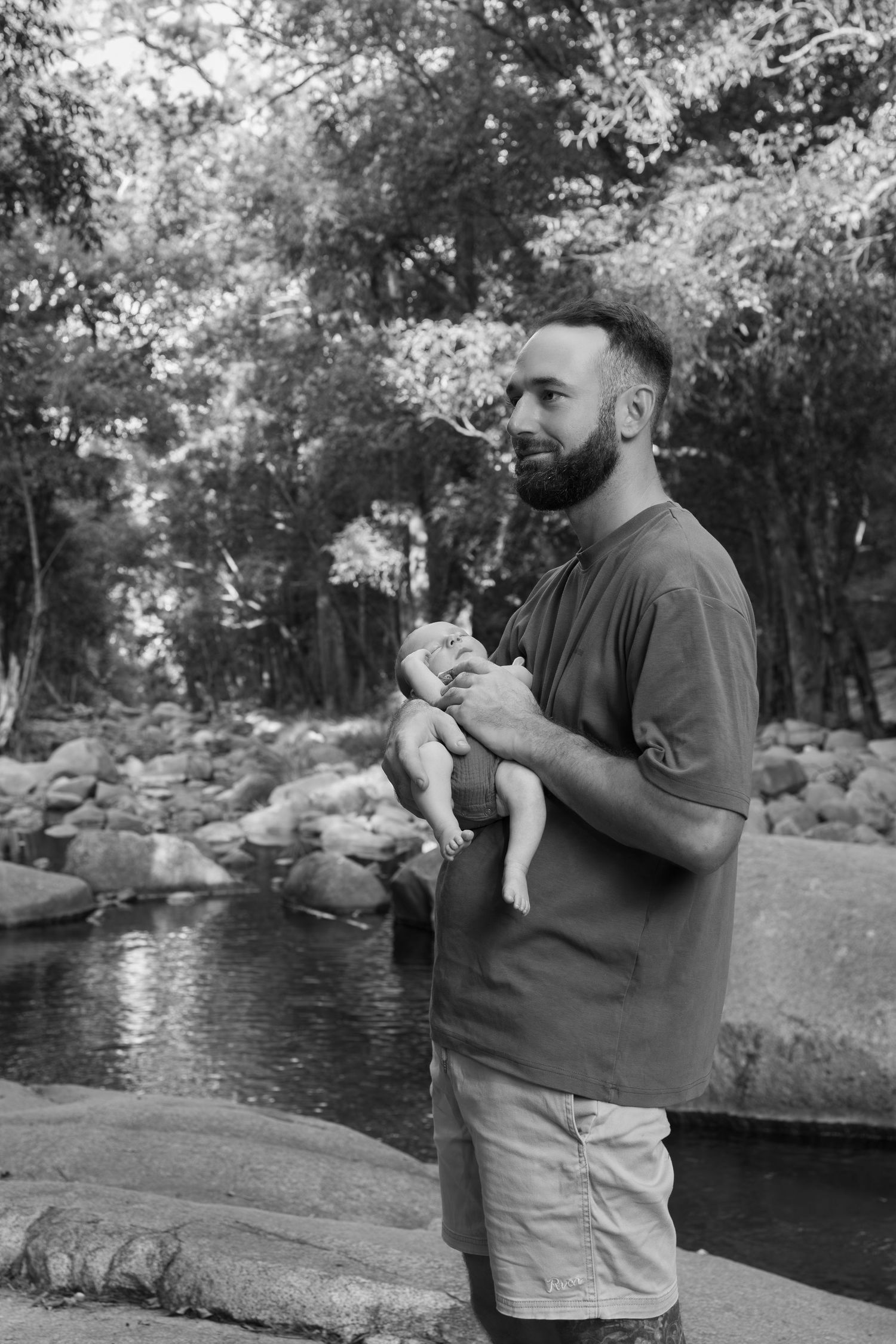 Bearded man holding a newborn baby outdoors near a rocky stream surrounded by lush green trees, black and white photo.