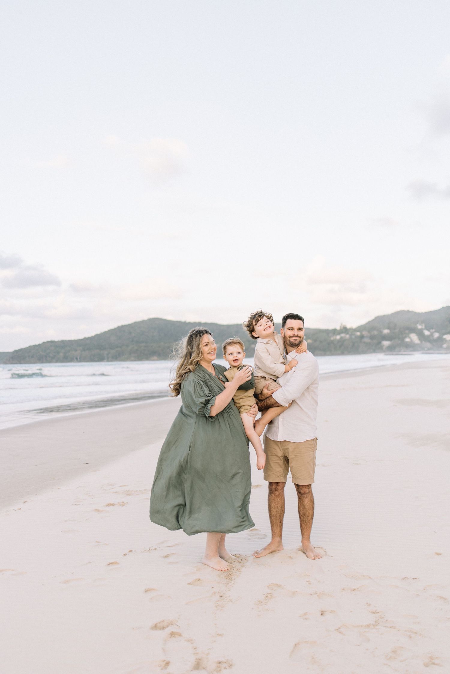 Family of four posing on a sandy beach at sunset, parents holding two young children, hills visible in the background.