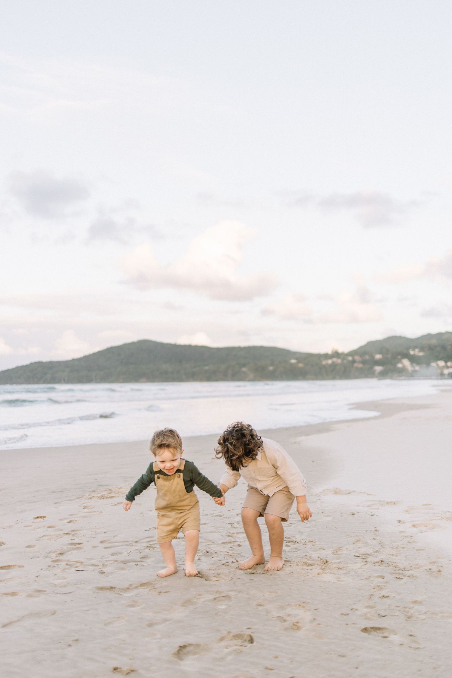 Two young children hold hands and play together on a sandy beach at sunset.