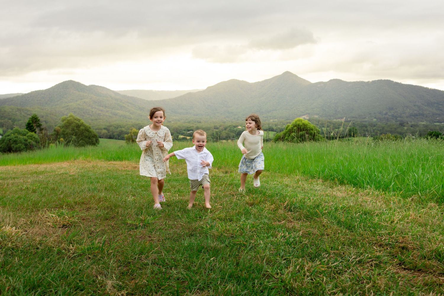 Three children running and playing in a lush green field with scenic mountain backdrop on a cloudy day.