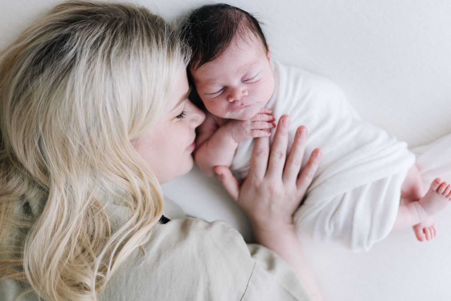A blonde mother tenderly kisses her sleeping newborn baby wrapped in white, captured from above.