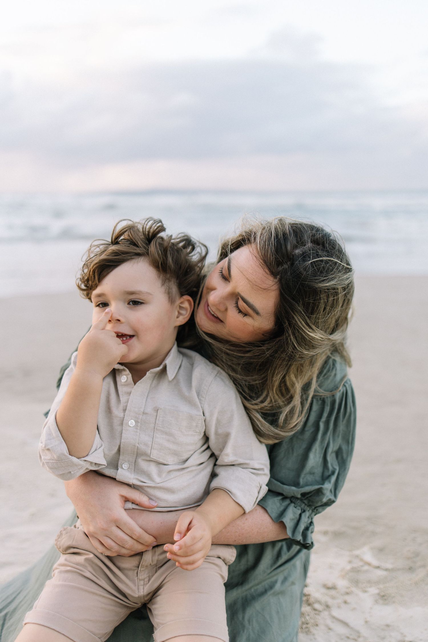 A smiling mother cuddles her young son on a sandy beach with ocean waves in the background.