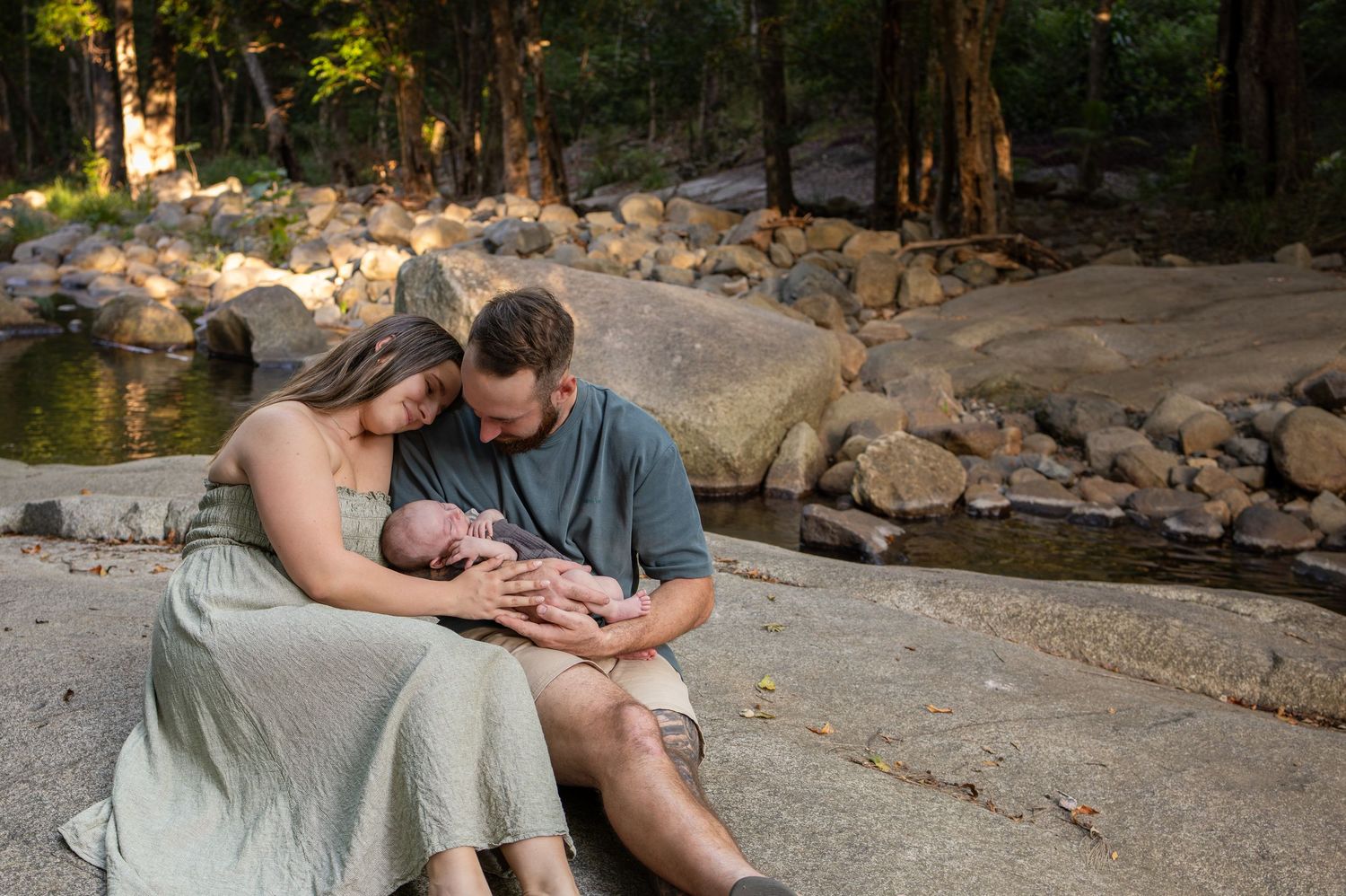 New parents sit on rocks by a forest stream, lovingly cradling their newborn baby in a serene outdoor setting.